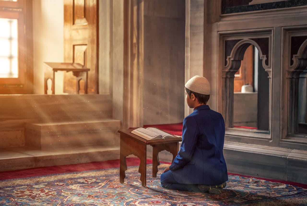 about-01 A young boy kneels in prayer inside a mosque with sunlight streaming through the window.
