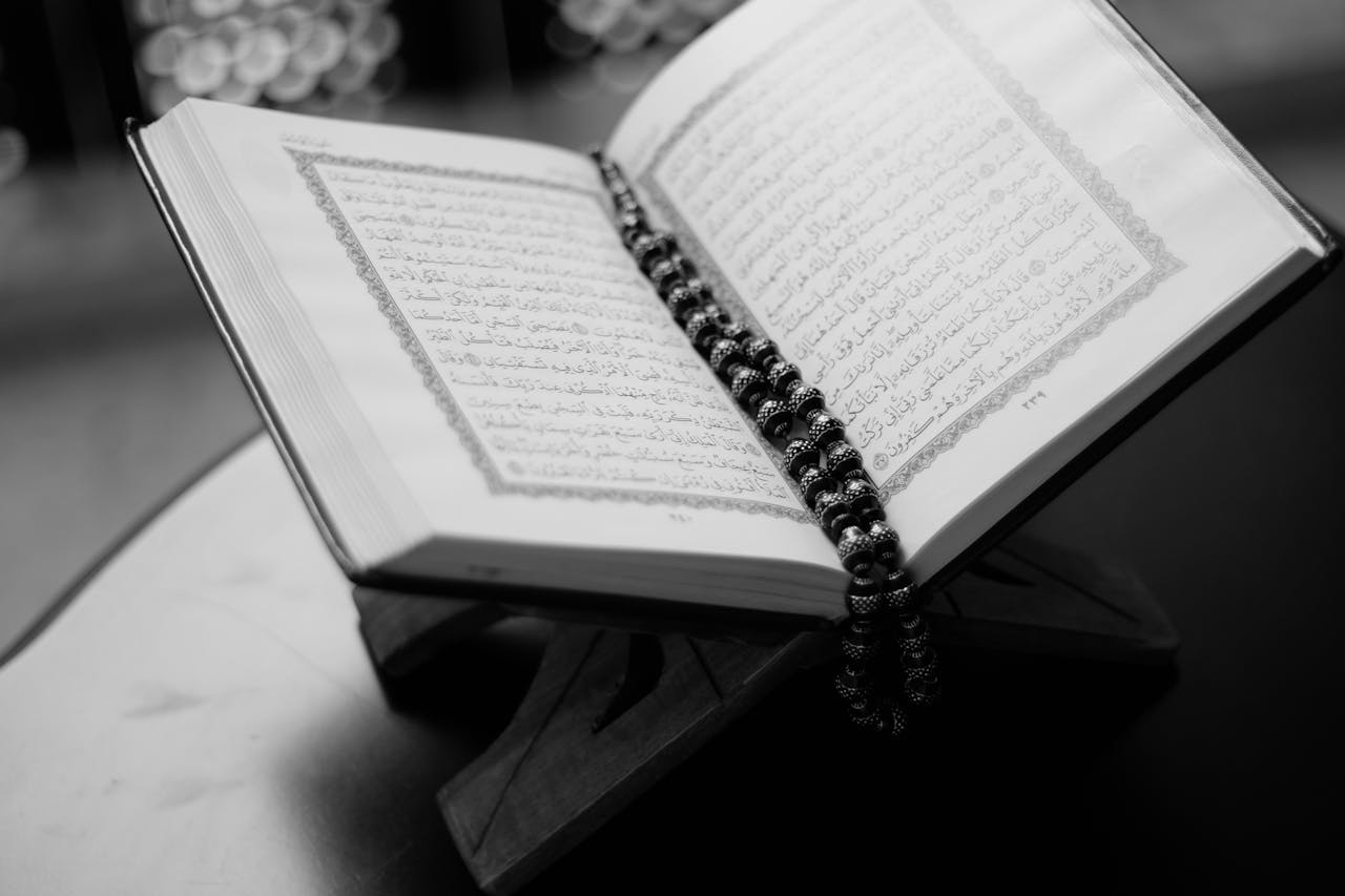 our-services-2 A black and white close-up of an open Quran on a wooden stand with prayer beads, symbolizing Islamic faith and devotion.